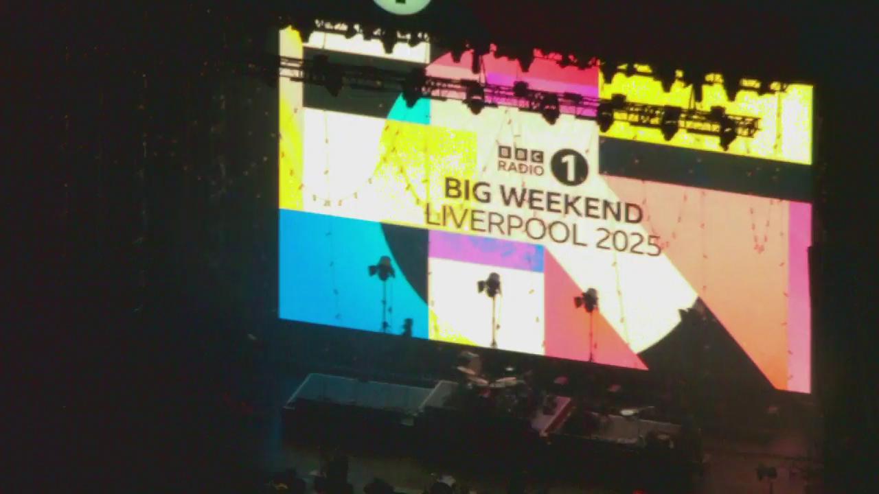 A large screen displays the BBC Radio 1 logo and the text 'BIG WEEKEND LIVERPOOL 2025' against a colorful geometric background. The stage below is set up with musical instruments and equipment, indicating preparation for a live event or concert.