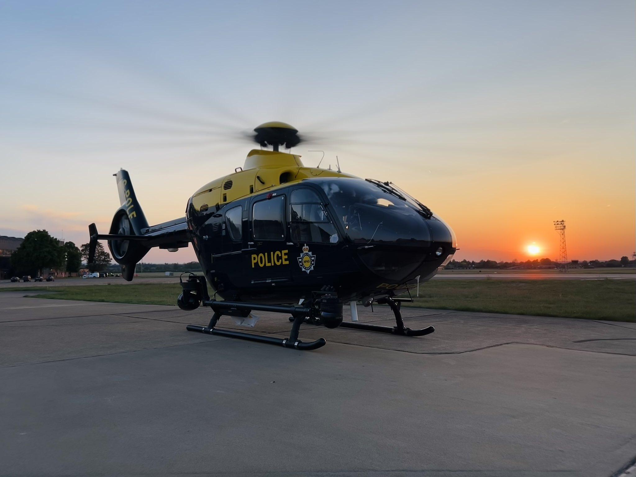 A police helicopter is parked on a tarmac at sunset. The helicopter is black with yellow accents and has the word 'POLICE' and a police badge emblem on its side. The rotors are spinning, and the sun is low on the horizon, casting an orange glow across the sky. There are buildings and trees in the background, as well as a tall light tower to the right of the image.