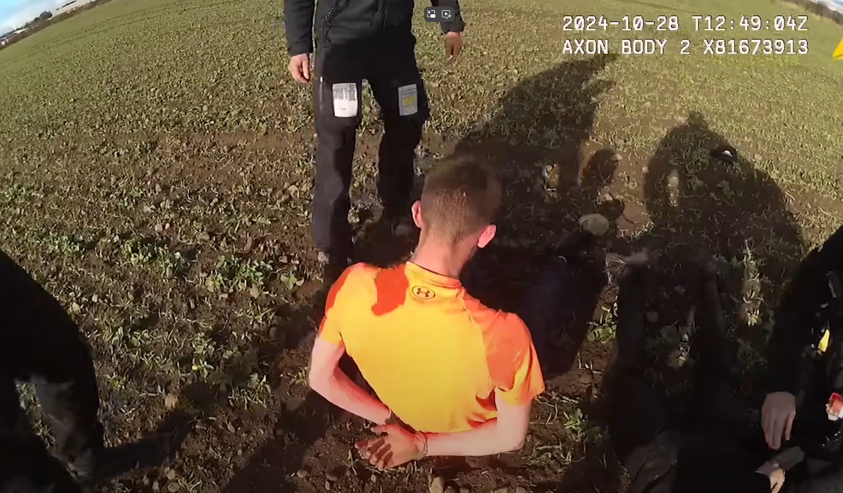 A person in an orange shirt is seated on the ground with hands cuffed behind their back, surrounded by at least three police officers. The image is from body-worn camera footage dated 28 October 2024.