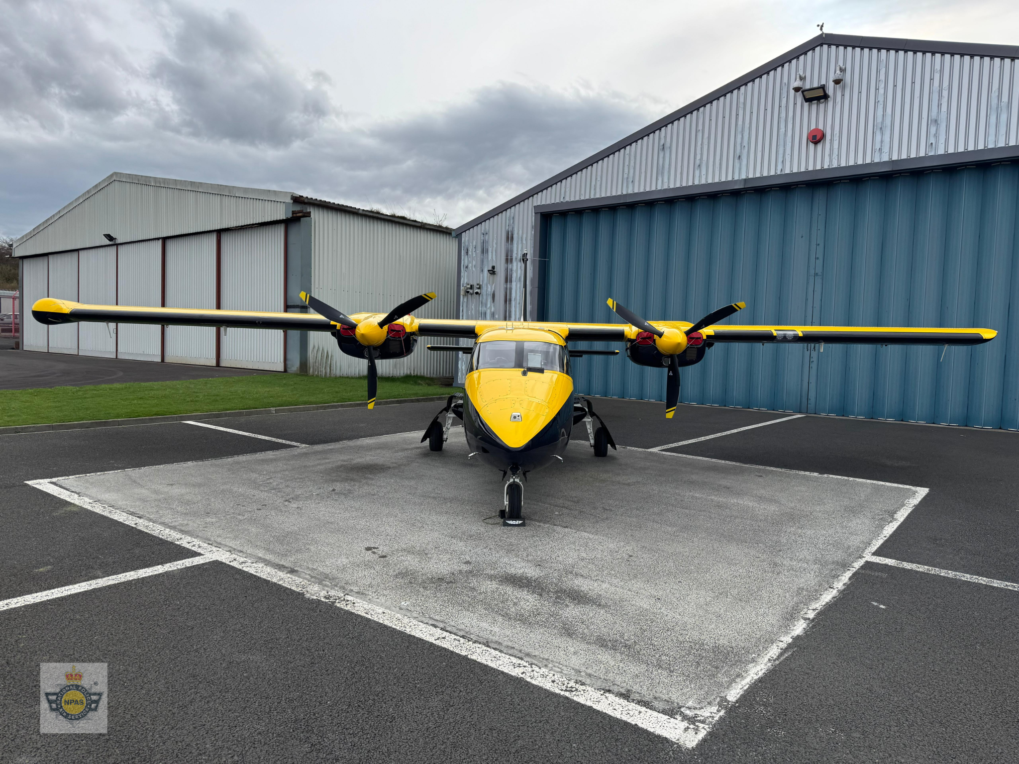 A yellow and black twin-engine propeller aircraft is parked on a tarmac in front of a large blue hangar. The plane has a high-wing design with a propeller on each wing. In the background, there’s another hangar with red and white walls under a cloudy sky.