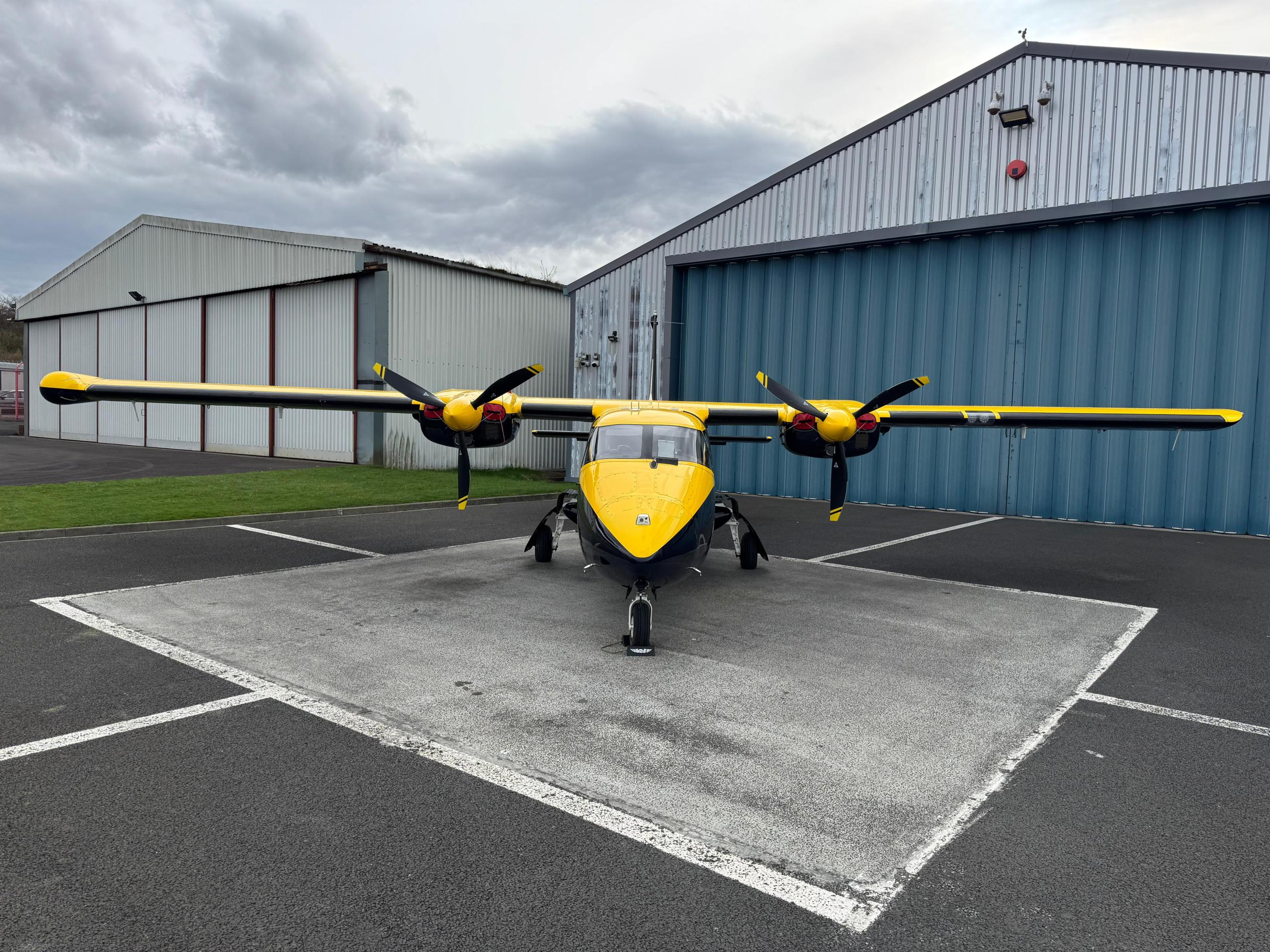 A yellow and black twin-engine propeller aircraft is parked on a tarmac in front of a large blue hangar. The plane has a high-wing design with a propeller on each wing. In the background, there’s another hangar with red and white walls under a cloudy sky.