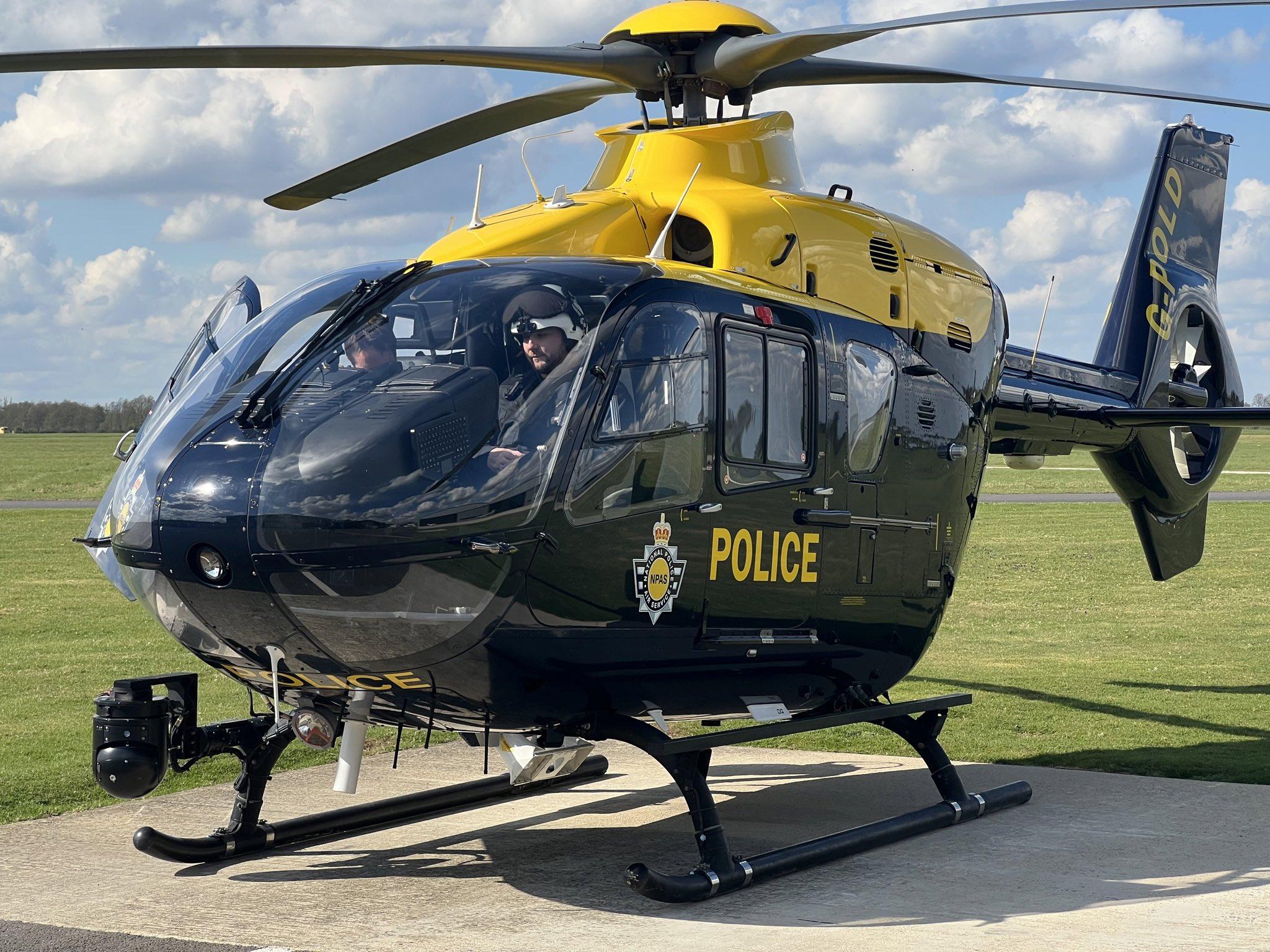 A yellow and black NPAS police helicopter with the registration code G-POLD parked on a concrete surface. The word 'POLICE' and the NPAS emblem are visible on the side. Two people are seated in the cockpit, with their faces obscured. Grass and a partly cloudy sky form the background.