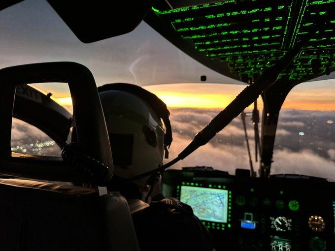 Pilot in a helicopter cockpit at sunset, with glowing instruments and a view above the clouds.