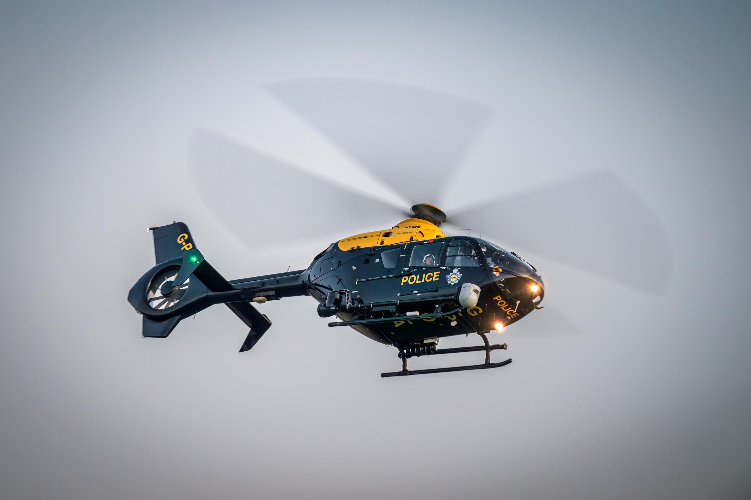 A black and yellow police helicopter in flight with spinning rotor blades, displaying the word 'POLICE' and a badge emblem on its side. Lights are visible on the tail and front of the aircraft.