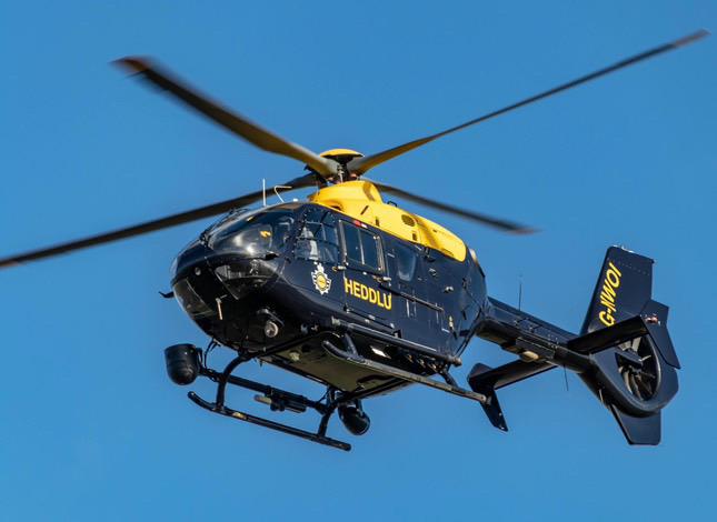 A police helicopter with the word "HEDDLU" written on its side is flying in a clear blue sky. The helicopter has a yellow and dark blue color scheme, with the registration number "G-WONI" visible on the tail.