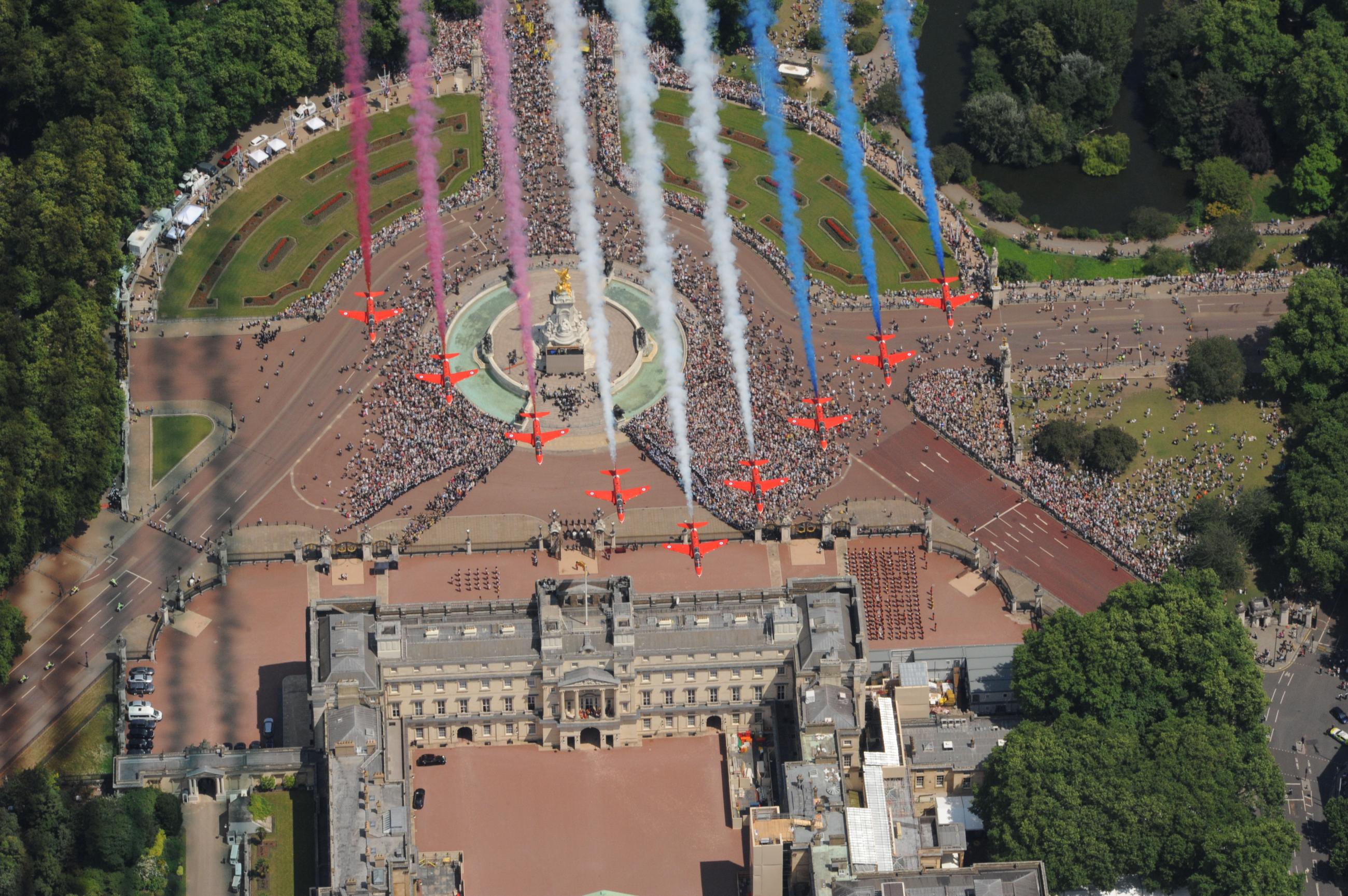 Aerial view of Buckingham Palace with a large crowd gathered in front. Nine aircraft fly in formation overhead, releasing red, white, and blue smoke trails during a celebratory event