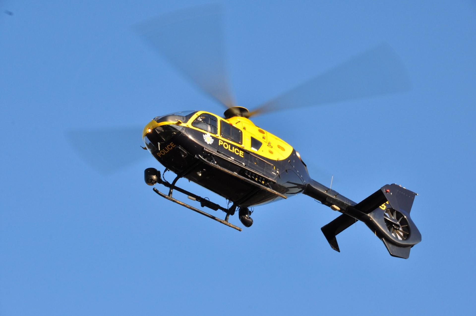 A blue and yellow police helicopter in flight against a clear blue sky, with the word 'POLICE' visible on its side and rotor blades blurred in motion
