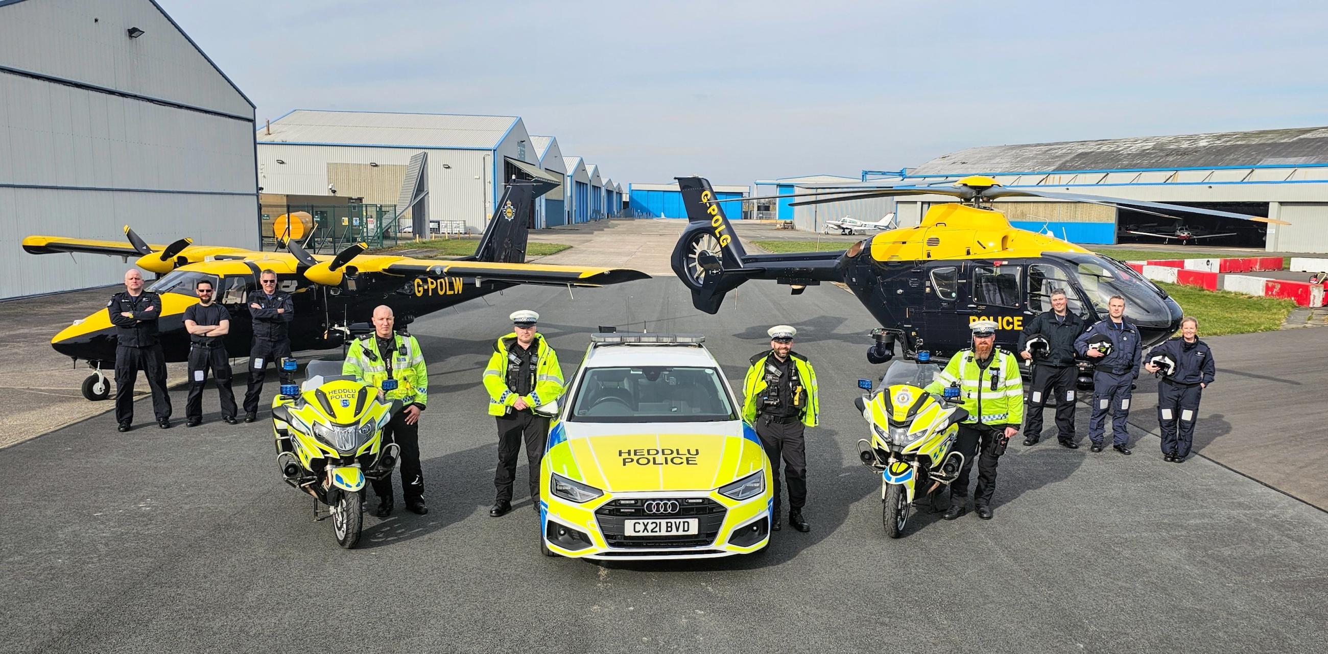 A group of police officers stand in front of two police motorcycles, a police car labeled 'HEDDLU POLICE' with the license plate 'CX21 BVD', and two aircraft—a plane marked 'G-POLW' and a helicopter labelled 'POLICE'—at an airfield