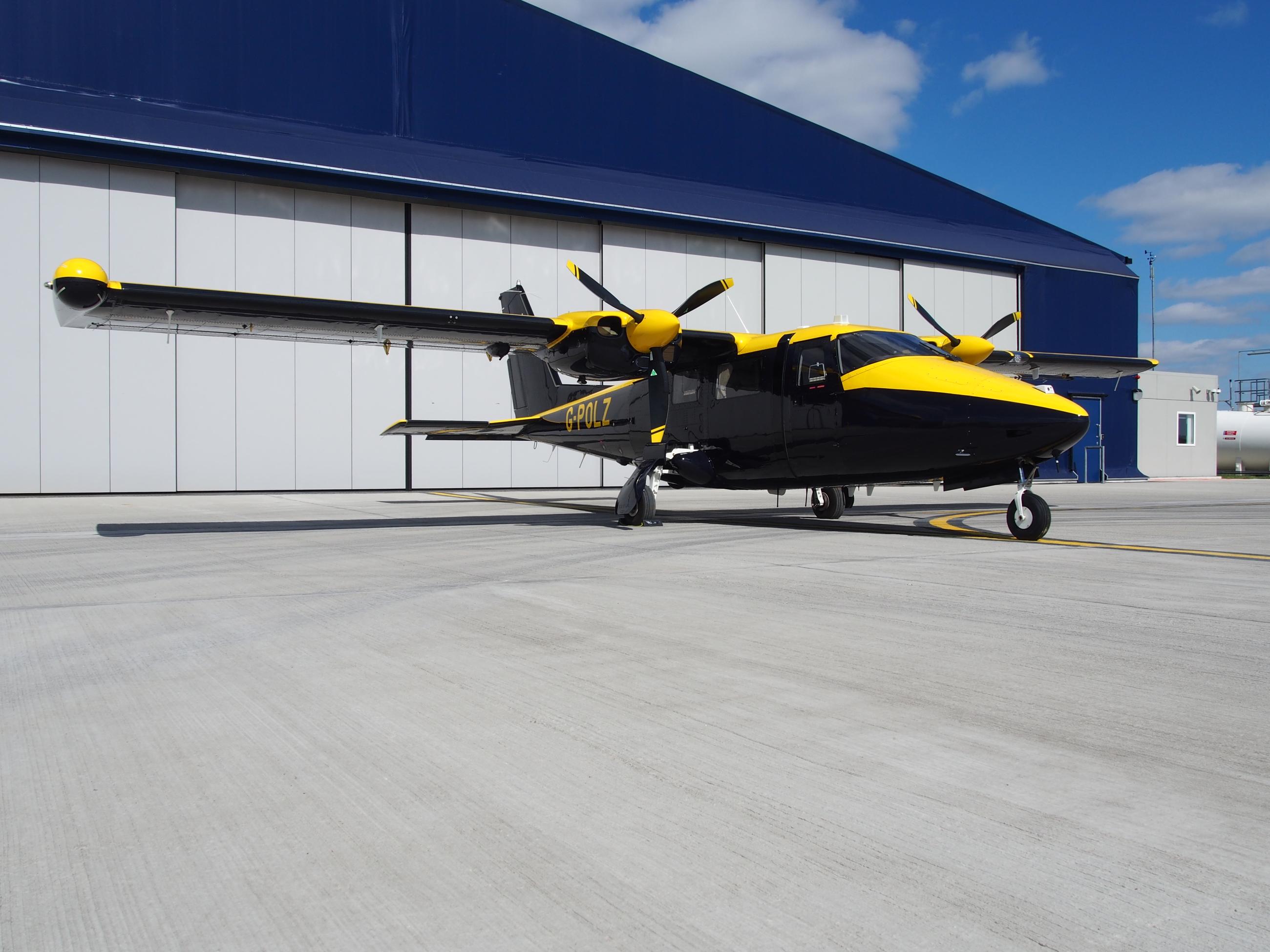 Twin-engine NPAS aircraft with black and yellow livery, registration G-POLZ, parked on an airport tarmac in front of a large blue hangar.