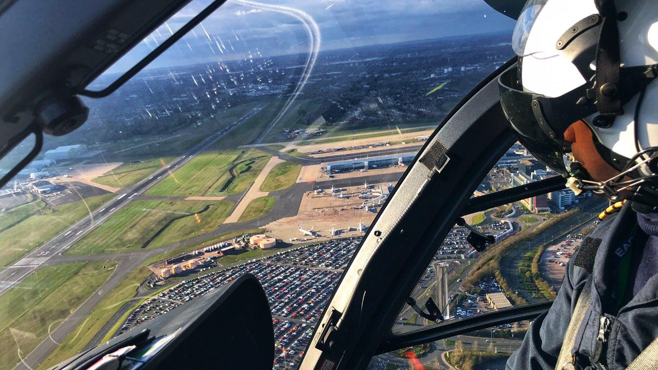 View from inside a helicopter cockpit overlooking an airport. The pilot, wearing a helmet and headset, is visible on the right. Below are runways, taxiways, airport buildings, and a large parking lot. A cityscape is visible on the horizon under a partly cloudy sky.