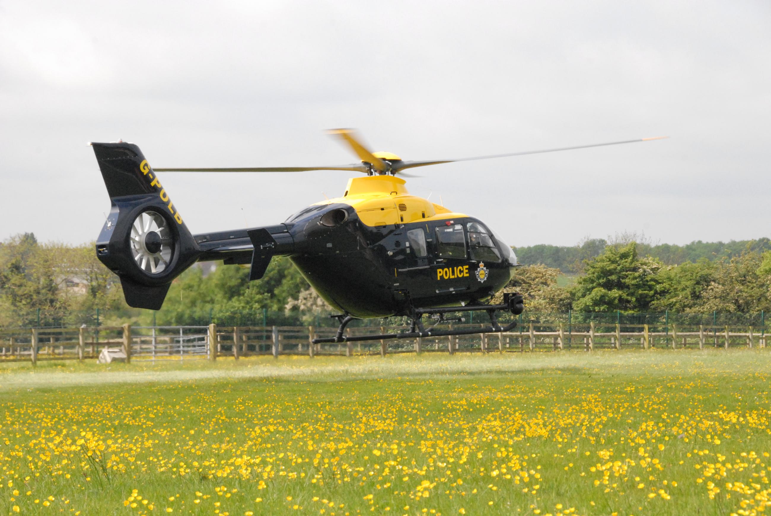 A black and yellow police helicopter with the tail number 'G-POLC' hovers above a field of yellow flowers. The word 'POLICE' and an emblem are visible on its side. In the background, there is a wooden fence, trees, and a cloudy sky.