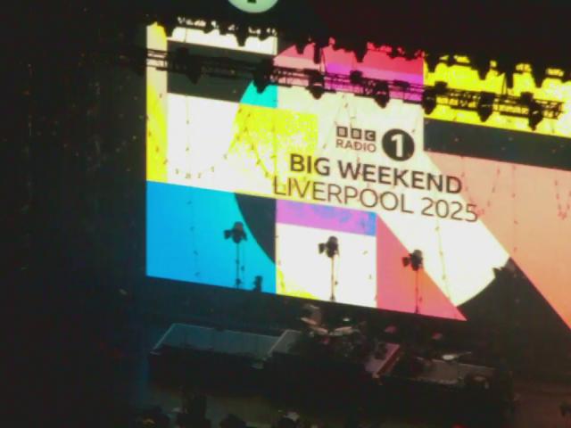A large screen displays the BBC Radio 1 logo and the text 'BIG WEEKEND LIVERPOOL 2025' against a colorful geometric background. The stage below is set up with musical instruments and equipment, indicating preparation for a live event or concert.