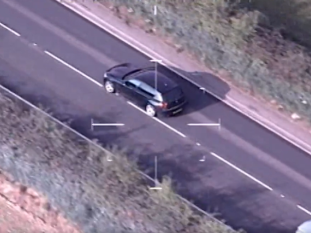 An aerial view of a black car driving on a two-lane road with greenery on both sides. The image appears to be taken from a police helicopter or drone, as indicated by the Gloucestershire Constabulary logo in the top left corner and crosshairs centred on the car.