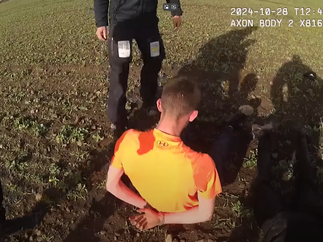 A person in an orange shirt is seated on the ground with hands cuffed behind their back, surrounded by at least three police officers. The image is from body-worn camera footage dated 28 October 2024.