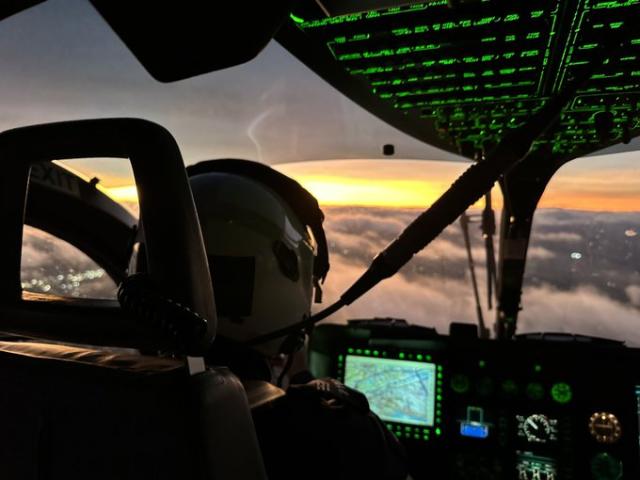 Pilot in a helicopter cockpit at sunset, with glowing instruments and a view above the clouds.