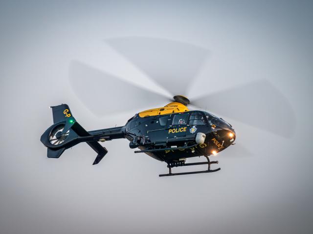 A black and yellow police helicopter in flight with spinning rotor blades, displaying the word 'POLICE' and a badge emblem on its side. Lights are visible on the tail and front of the aircraft.