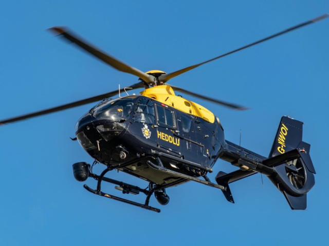 A police helicopter with the word "HEDDLU" written on its side is flying in a clear blue sky. The helicopter has a yellow and dark blue color scheme, with the registration number "G-WONI" visible on the tail.