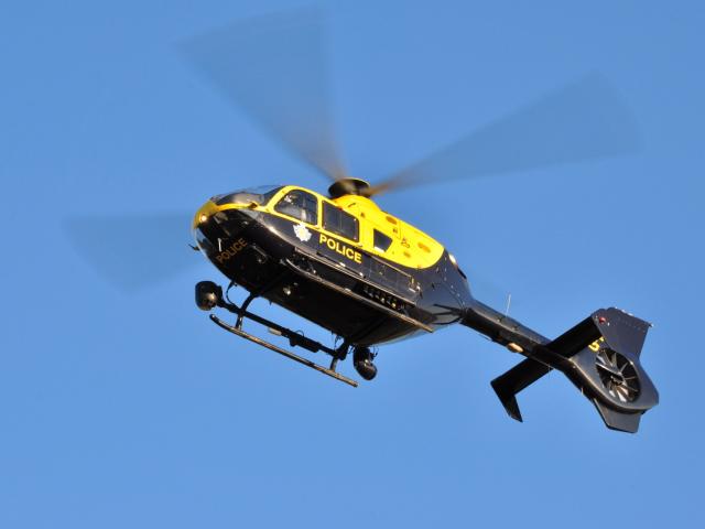 A blue and yellow police helicopter in flight against a clear blue sky, with the word 'POLICE' visible on its side and rotor blades blurred in motion