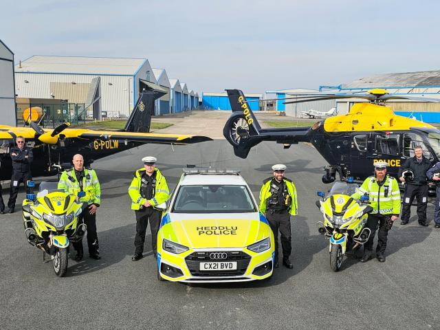A group of police officers stand in front of two police motorcycles, a police car labeled 'HEDDLU POLICE' with the license plate 'CX21 BVD', and two aircraft—a plane marked 'G-POLW' and a helicopter labelled 'POLICE'—at an airfield