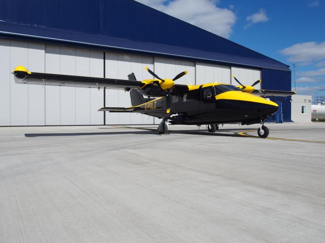 Twin-engine NPAS aircraft with black and yellow livery, registration G-POLZ, parked on an airport tarmac in front of a large blue hangar.