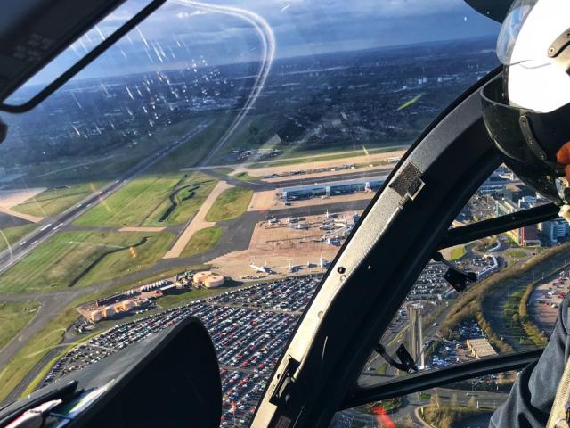 View from inside a helicopter cockpit overlooking an airport. The pilot, wearing a helmet and headset, is visible on the right. Below are runways, taxiways, airport buildings, and a large parking lot. A cityscape is visible on the horizon under a partly cloudy sky.