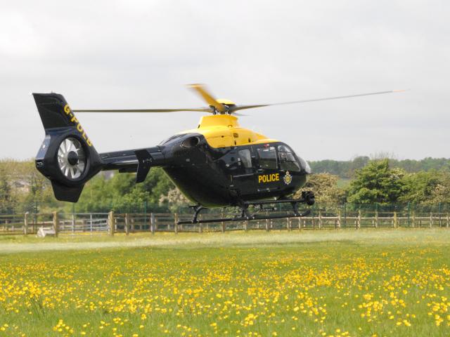 A black and yellow police helicopter with the tail number 'G-POLC' hovers above a field of yellow flowers. The word 'POLICE' and an emblem are visible on its side. In the background, there is a wooden fence, trees, and a cloudy sky.